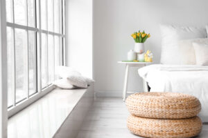 Janela de um quarto, ambiente todo branco, cama com travesseiros, uma mesa de cabeceira, com plantas e um puff de cordas