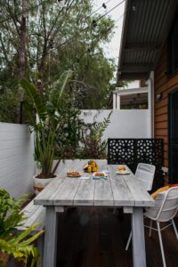 Jardim com mesa de me madeira cinza, plantas ao redor e comidas em cima.
